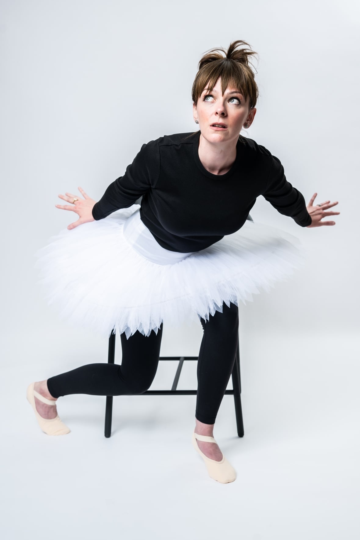 A woman with brown hair, a black body suit and a white tutu makes a lunging pose in front of a chair.
