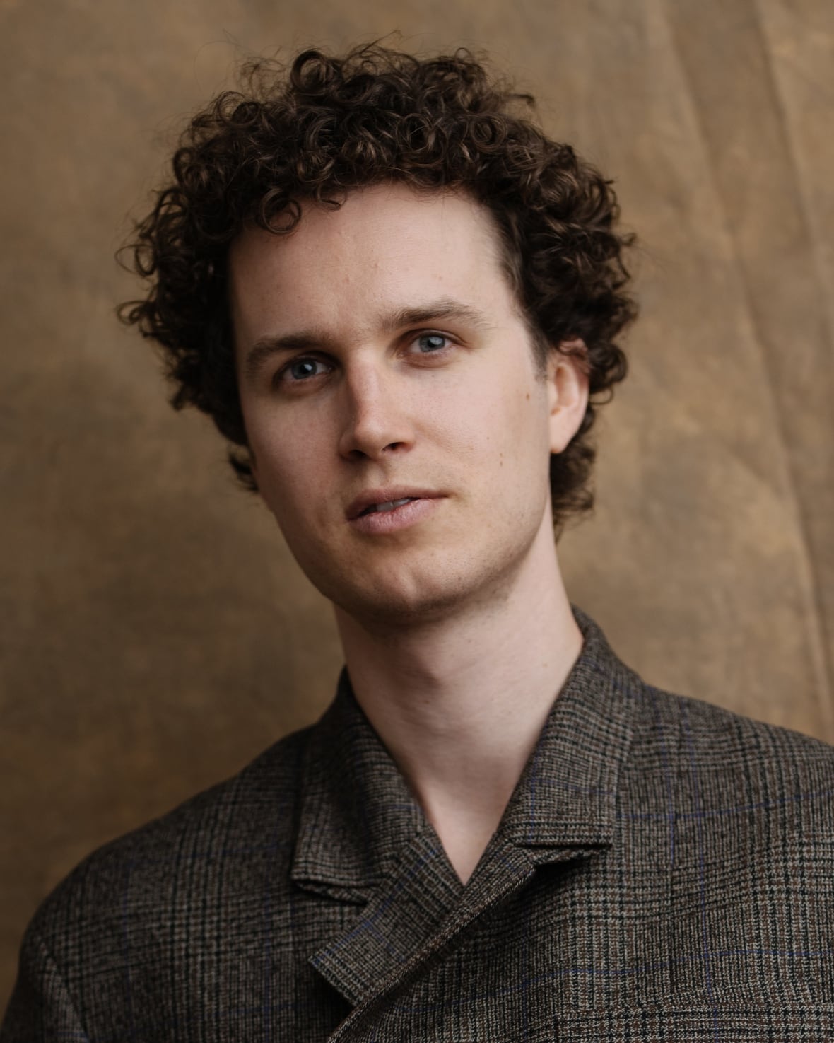 The headshot of a man with curly brown hair wearing a plaid patterned jacket.