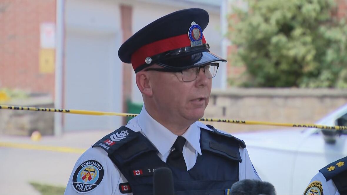 A middle-aged police officer speaks into reporters' microphones at a news conference outdoors on a city street on a sunny summer afternoon.
