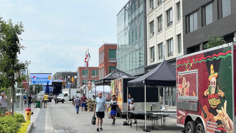 People walking through a downtown street with food trucks and tents for vendors. In the distance there's a stage