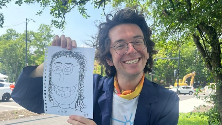 A young man with wavy black hair and glasses sits outside at a small table, covered in drawing supplies. He's smiling brightly and holding up a portrait of a bespectacled woman with long curly hair. It's crudely drawn in black marker, and one-dimensional, reminiscent of a child's drawing you might put on your fridge.