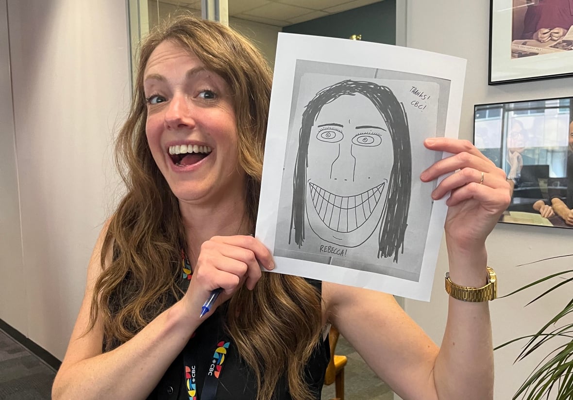 A woman with long, wavy, strawberry blonde hair stands in an office smiles as she holds up a print of a badly drawn portrait of herself with beady eyes, a large nose, and a maniacal toothy grin.