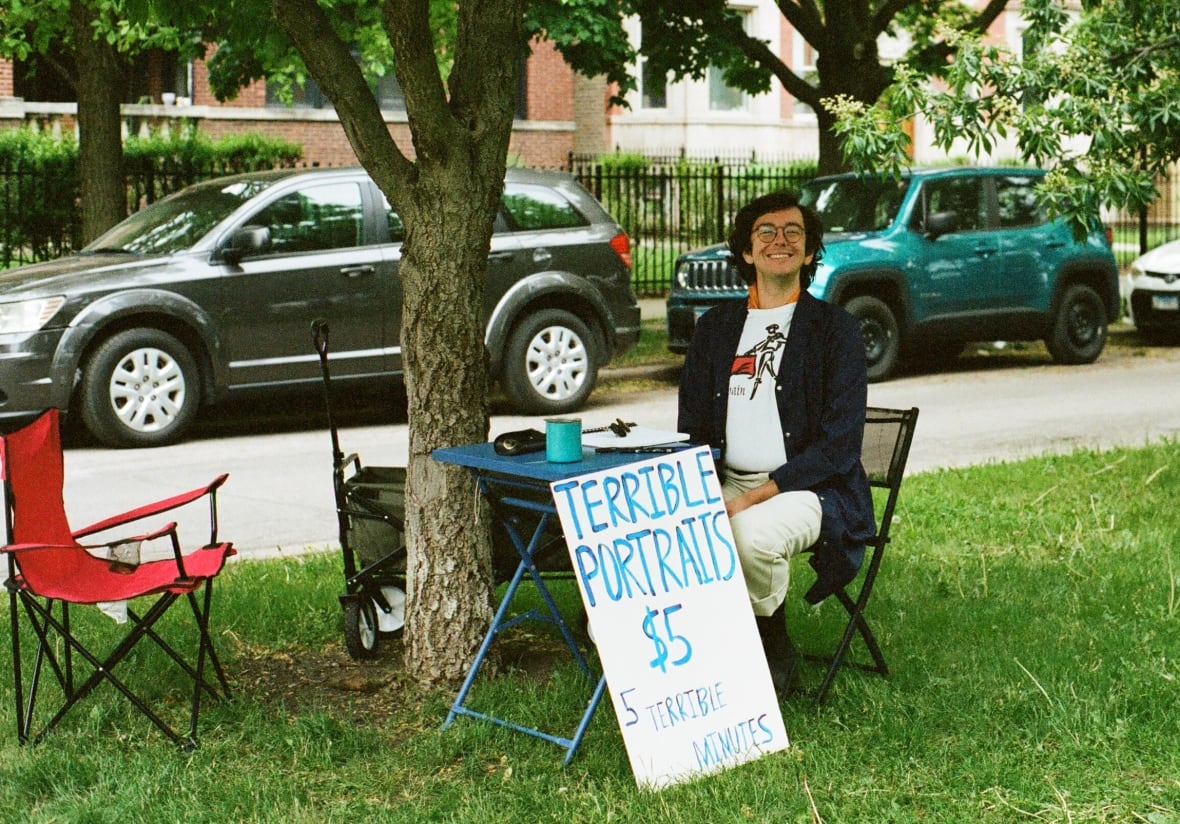 A young man with wavy black hair and glasses smiles brightly while sitting at a small folding table in the grass under a tree. Resting against the table is a hand-drawn sign, in blue, that reads: "Terrible portraits $5"