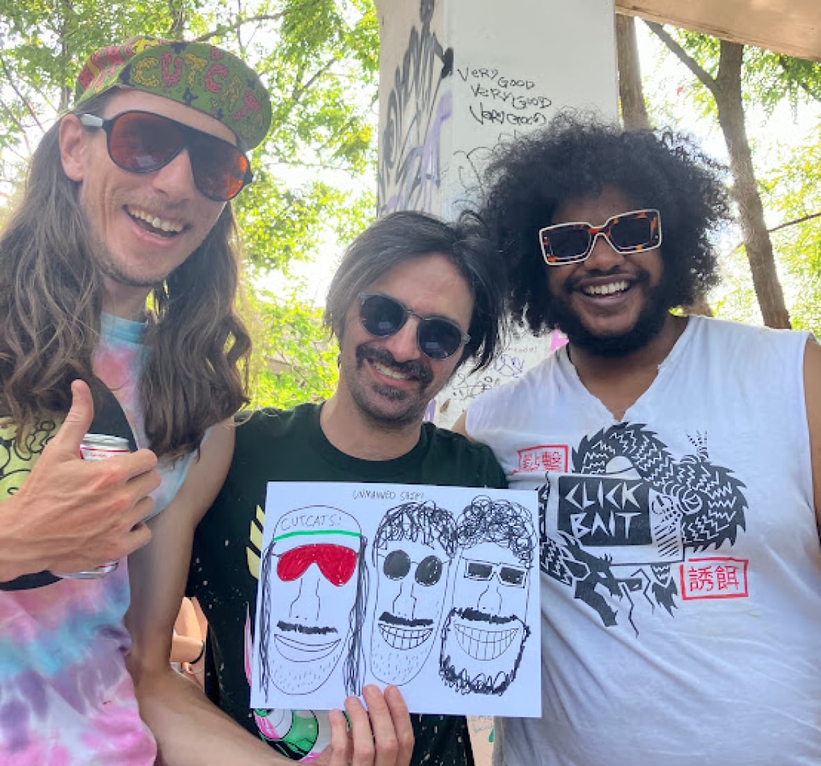 Three smiling men in sunglasses stand side-by-side under a graffiti-covered bridge and hold up a hand-drawn, poor-quality portrait of their grinning, mustachioed faces.
