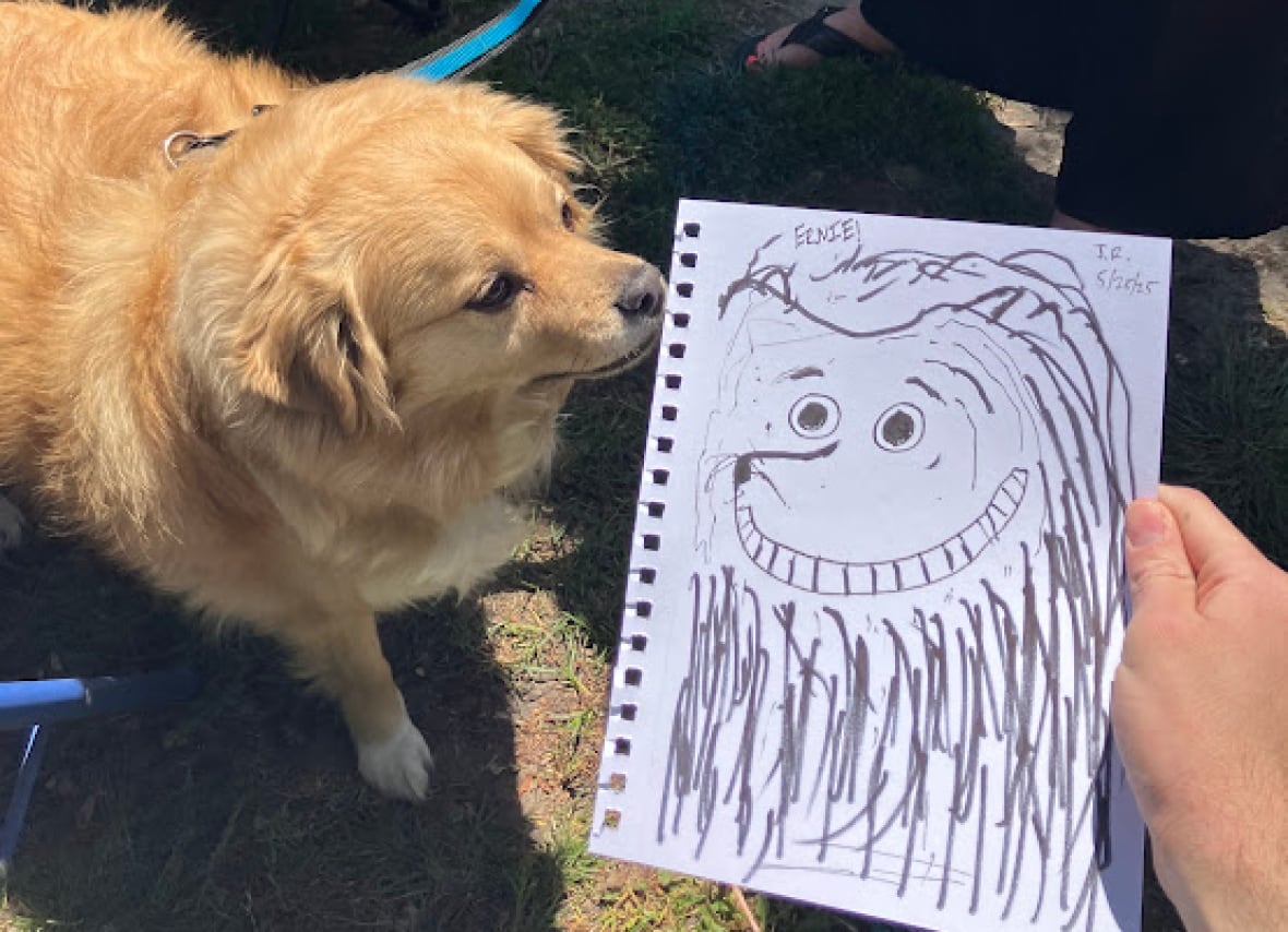 A small blonde dog, perhaps a Pomeranian or Pomeranian mix, sniffs a piece of paper with a terribly drawn sketch of itself with a toothy grin.