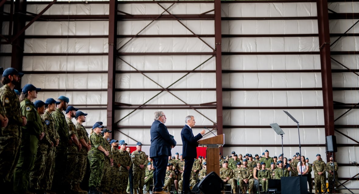 A man speaks at a podium in a hangar surrounded by soldiers.
