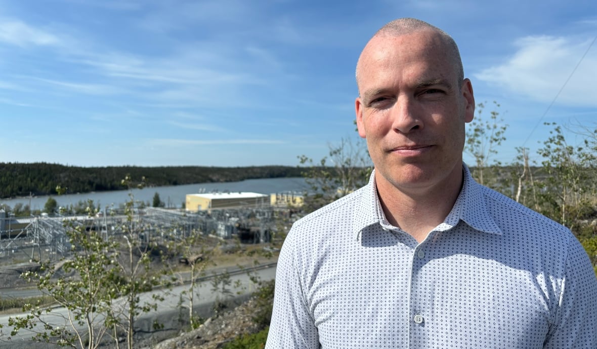 A man in a collared shirt stands outside on a sunny day. The Jackfish power plant is seen in the distance behind him.
