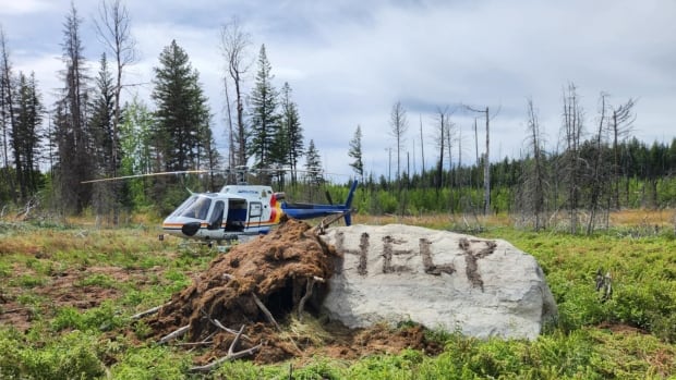 A rock smeared with dirt reading 'HELP' beside a small shelter made of sticks and mud and an RCMP helicopter in the background