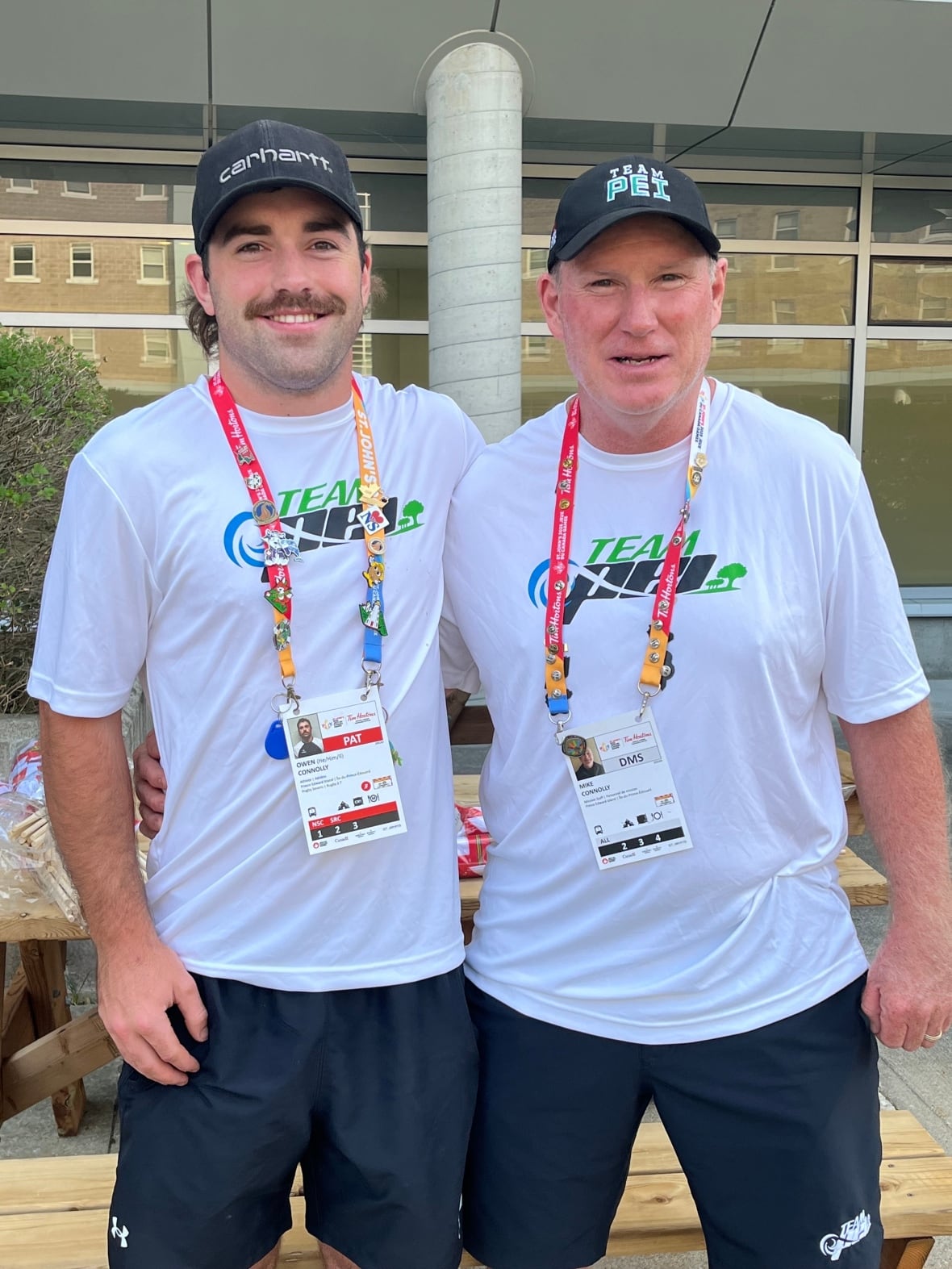 A younger man and an older man standing side by side and smiling at the camera. They are wearing shirts that read 'Team P.E.I.' 