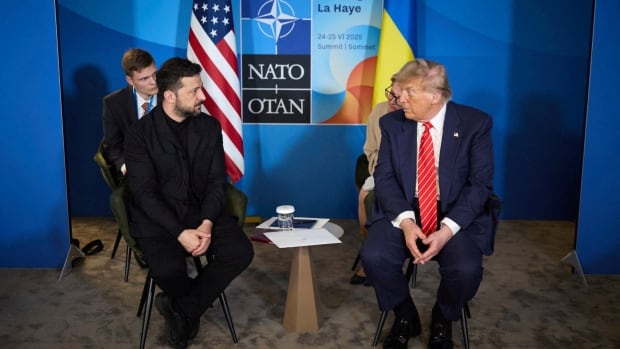 Two men sit in front of flags.