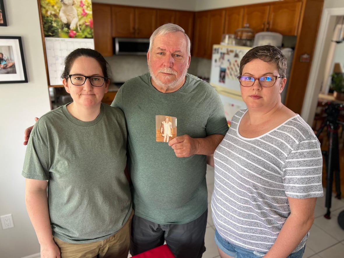 A man standing in the middle of his daughters. He is holding an old photo from the 1970s depicting a young woman. 