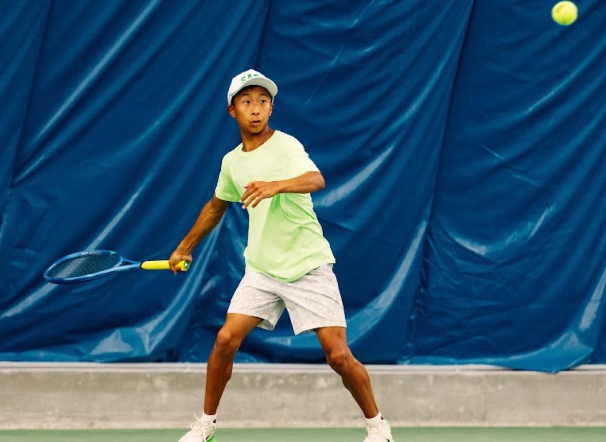 Young boy in green shirt looks at tennis ball in the midst of a match on an indoor court. 