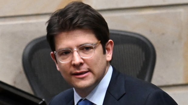 A bespectacled, cleanshaven and darkhaired main wearing a suit and tie is shown in closeup.