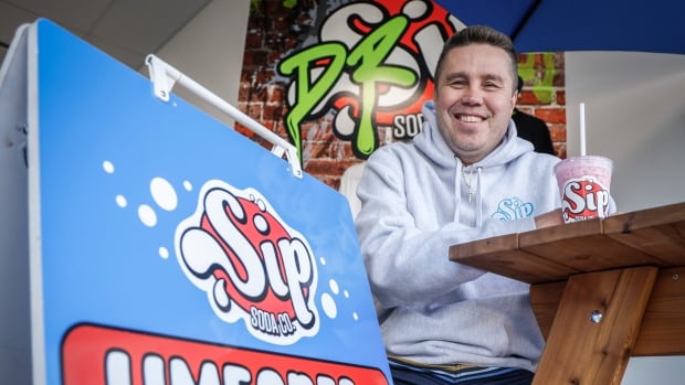 A man in a grey hoodie sits at a picnic table with a plastic soda cup. In the foreground, a red and blue sign reads "sip, limeades and lemonades." 