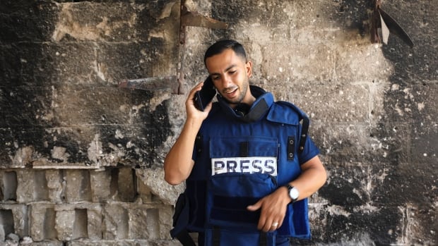 A smiling man in a blue "press" vest stands in front of a stone wall and speaks into a cellphone. 