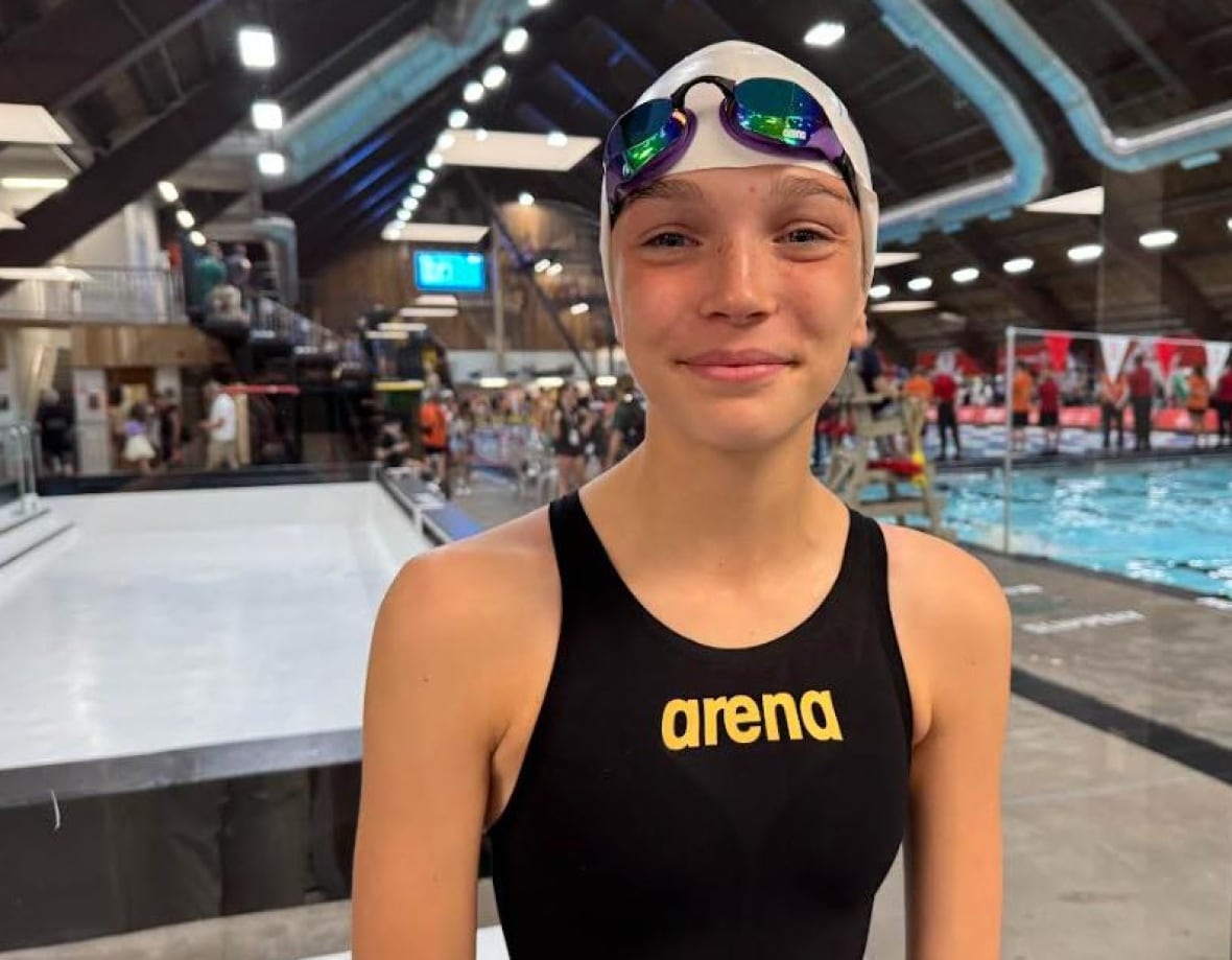 A girl in a swimsuit and swimcap stands beside a pool.