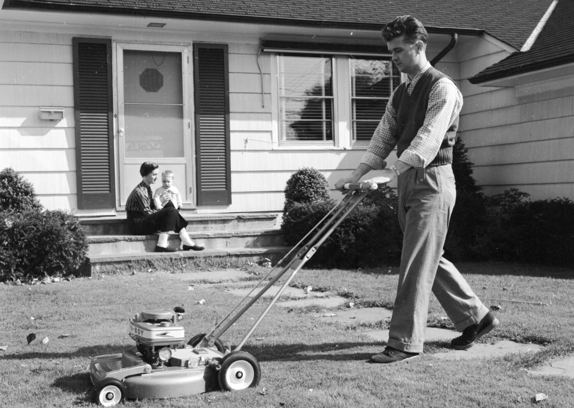A black and white archive photo of a man mowing a lawn in front of a house. A woman and baby sit on the front step