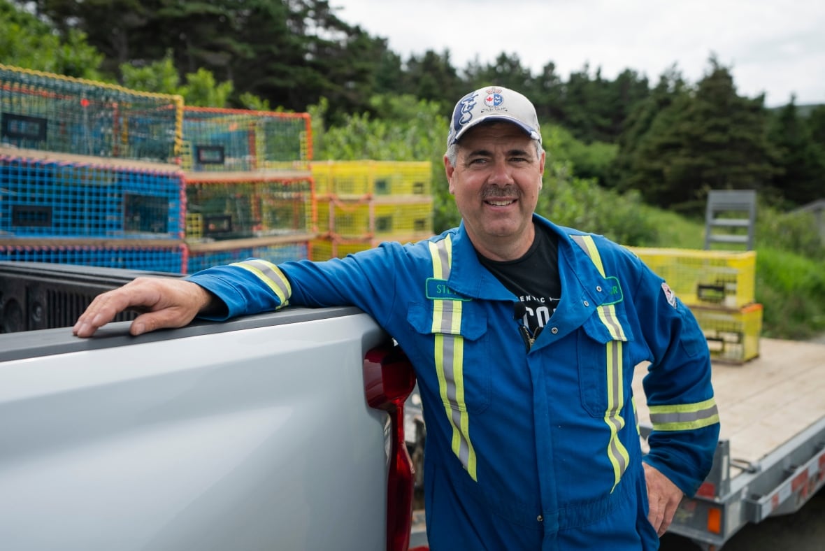 A man in a blue work outfir and greay baseball cap leans against a truck.