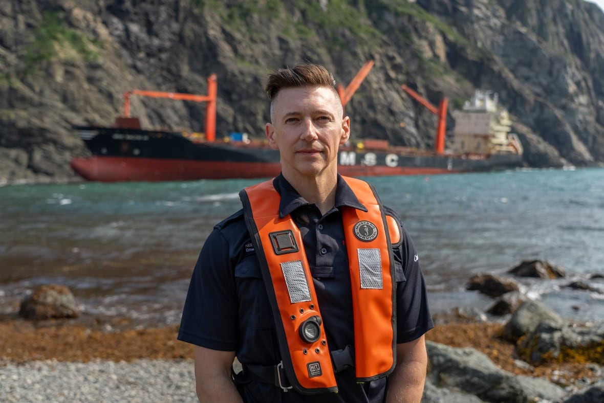 A man in a black uniform and an orange life preserver stands in front of a body of water with a large boat in the background.