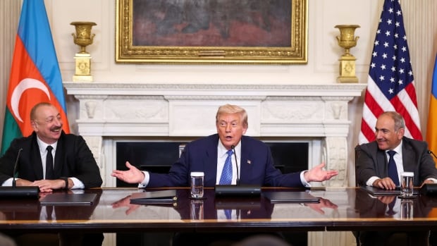 Three men sit at a large table with flags behind them.
