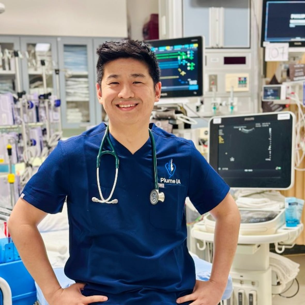 Man in scrubs with a stethoscope around his neck, hospital equipment behind him.