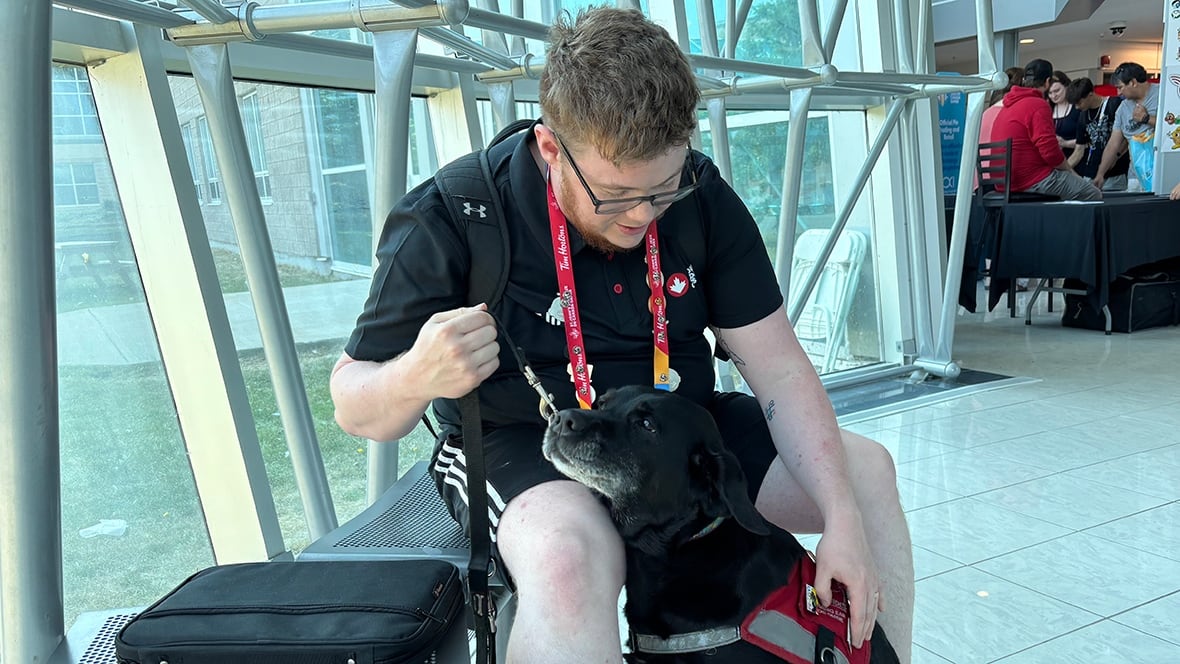 A person with glasses, a black short-leeved short and shorts, hold the leash of a black Labrador dog wearing a red service vest, who is sitting between the person's legs.
