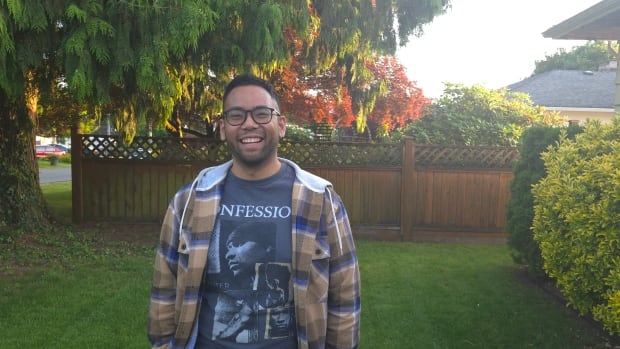 A man wearing a plaid shirt over a t-shirt stands in a yard smiling with a flowering tree and wooden fence behind him. 