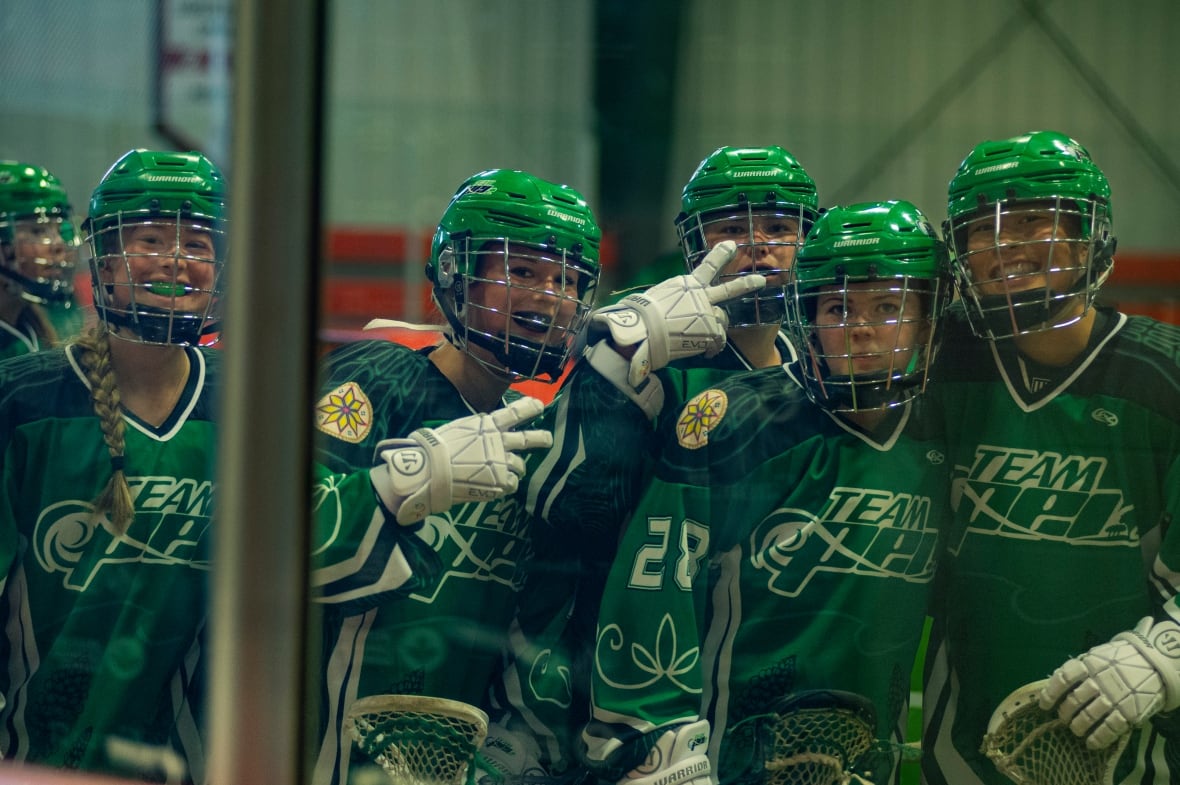 Six women's box lacrosse teammates, all wearing green jerseys and green helmets, smile for a photo.