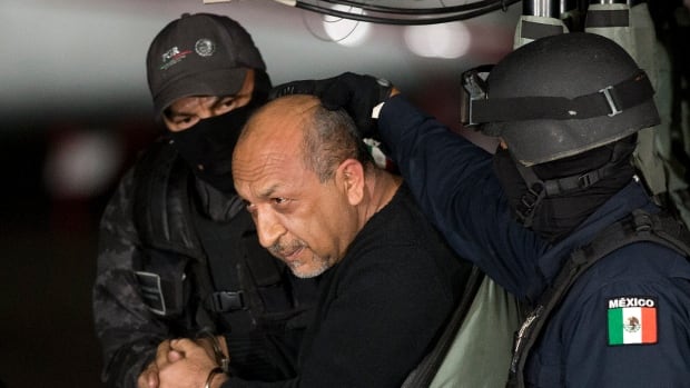 A balding man with a goattee is shown handcuffed and leaned over, seated in a vehicle.