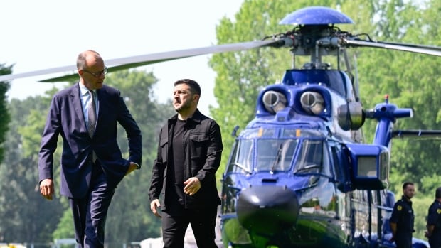 Two men walk near a helicopter. One is bearded and in all dark clothing, while the other is bespectacled, balding and cleanshaven, wearing a suit and tie.