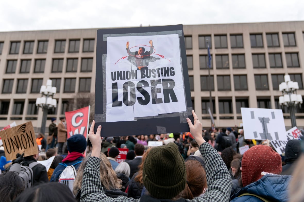 People who suffer from their appearance to the camera keep signals and paintings while facing a building in an outdoor protest. One of the prominent signs says: