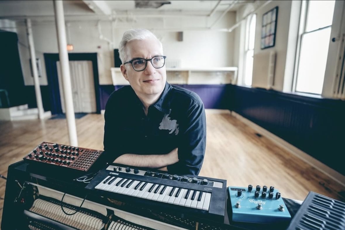 A white haired man with dark rimmed glasses leans on his piano keyboard in a studio.