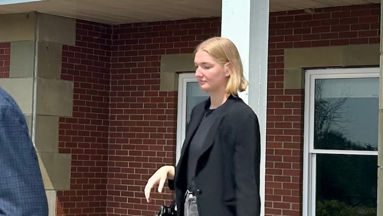 A young woman with a blonde bob wearing a black blazer and grey jeans walks out of a building not looking at the camera. 