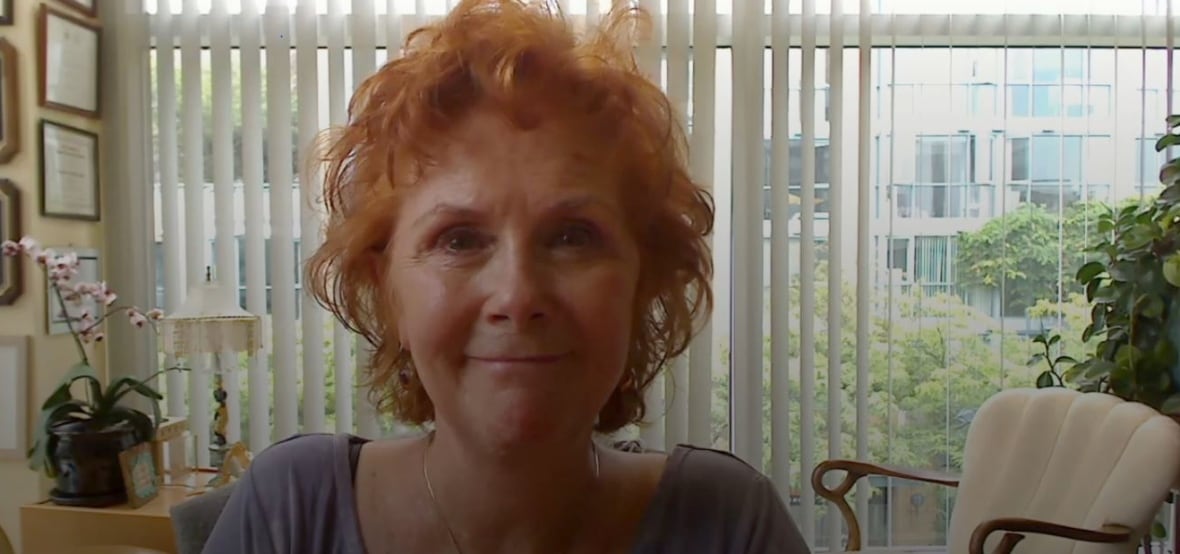 Una donna con i capelli corti e ricci, rossi, sorride con delle tende bianche, dei fiori rosa e una sedia imbottita bianca dietro di lei.