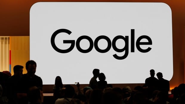 Audience members gather at Made By Google for new product announcements at Google on Tuesday, Aug. 13, 2024, in Mountain View, Calif. A large white screen with the word "Google" on it is shown with a few people silhouetted in black in front of it.