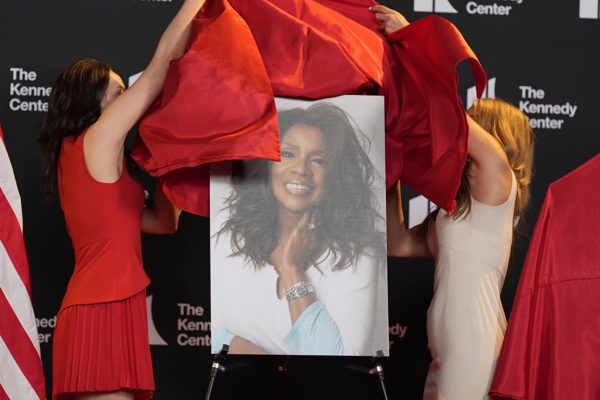 Zwei Frauen in Kleidern auf einem Stand nehmen Vorhänge von einem Porträt einer anderen Frau ab.