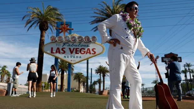 An Elvis impersonator with a guitar stands near a sign that reads: Welcome to Fabulous Las Vegas Nevada, which is flanked by two palm trees. Several tourists stand in front of the sign making cellphone stills.
