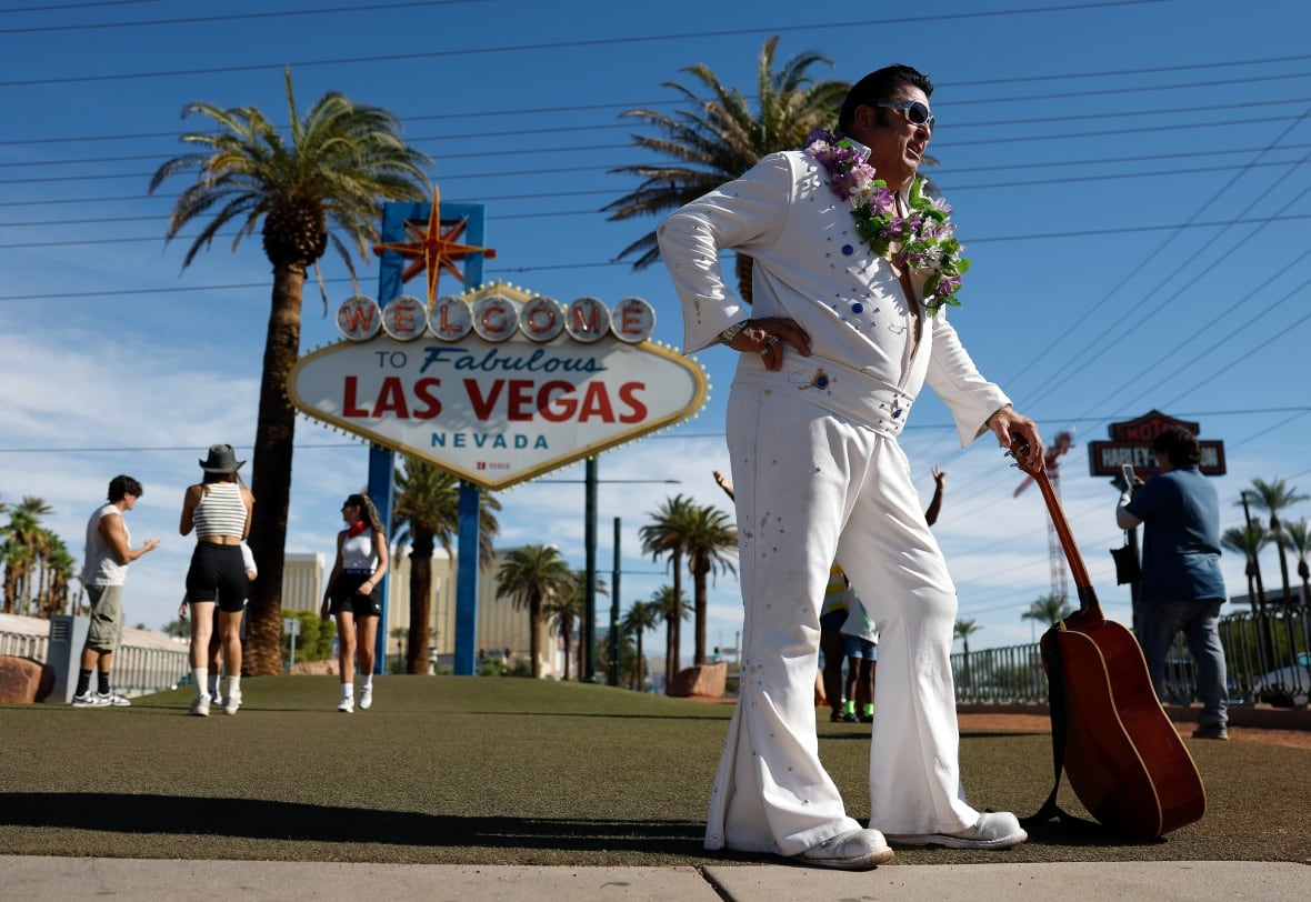 An Elvis impersonator with a guitar stands near a sign that reads: Welcome to Fabulous Las Vegas Nevada, which is flanked by two palm trees. Several tourists stand in front of the sign making cellphone stills.