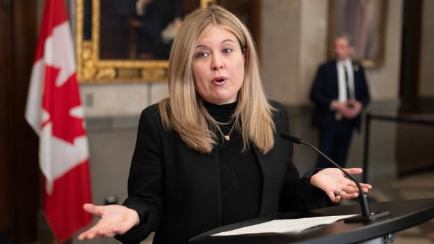 Conservative MP Michelle Rempel Garner speaks from the podium wearing a black suit, her arms outstretches either side.