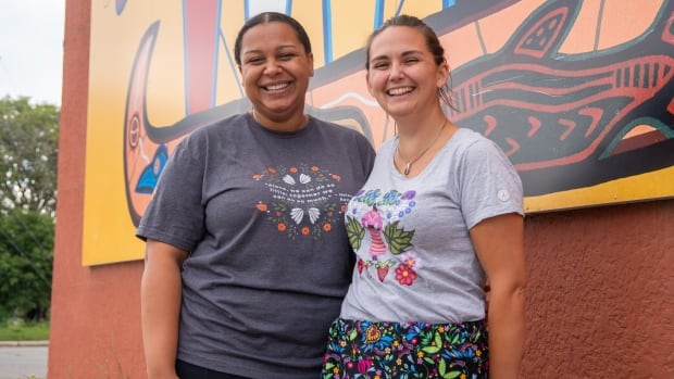 Ashley Wilkinson, left, and Champagne Thomson are seen outside of Shelter House in Thunder Bay, Ont., where Wilkinson has been interviewing Black and Indigenous clients about their experiences with racism in health-care settings. Thomson is the shelter's development manager. Two people are seen standing outside a building in front of a painted mural.