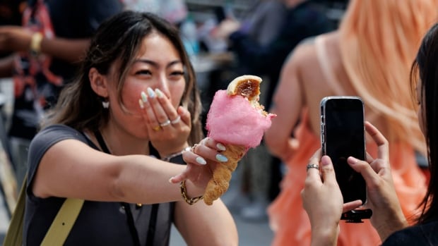Woman holds a cotton candy wrapped butter tart towards another woman holding a phone.