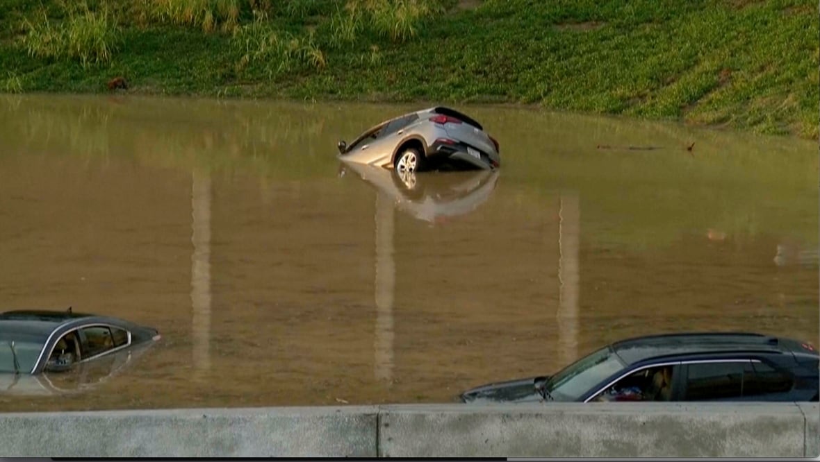 Parts of three vehicles are represented strongly overwhelmed in brown water near a grassy hill.
