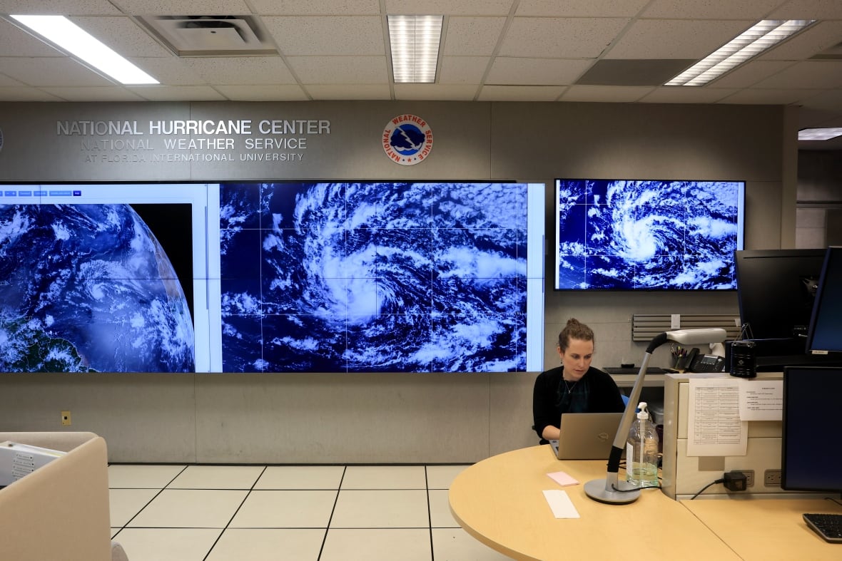 A woman works at a computer with monitors showing hurricanes behind her. 