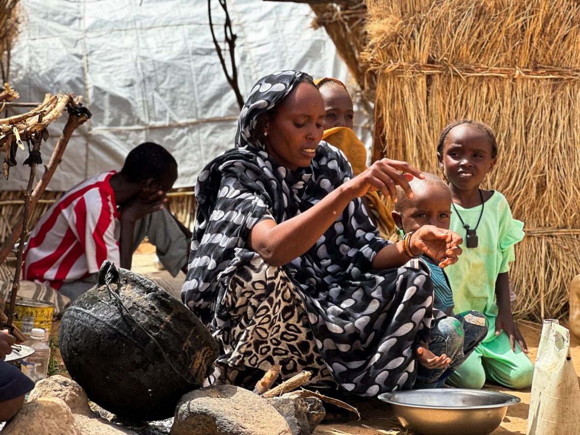 A woman wears a long black and white dress sitting on the floor, spraying the ingredients in a metal bowl and gathering children around it.