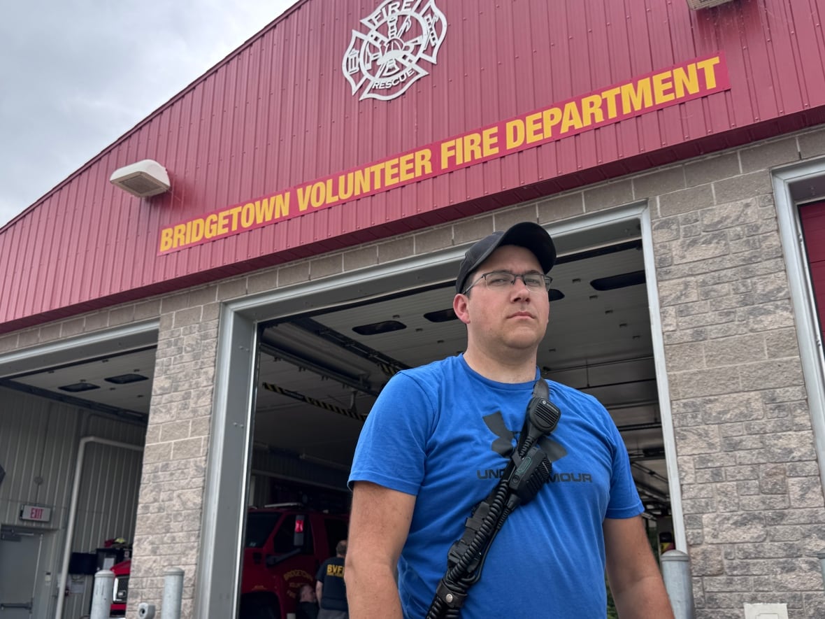 A man in a blue t shirt and glasses stands in front of a fire station.