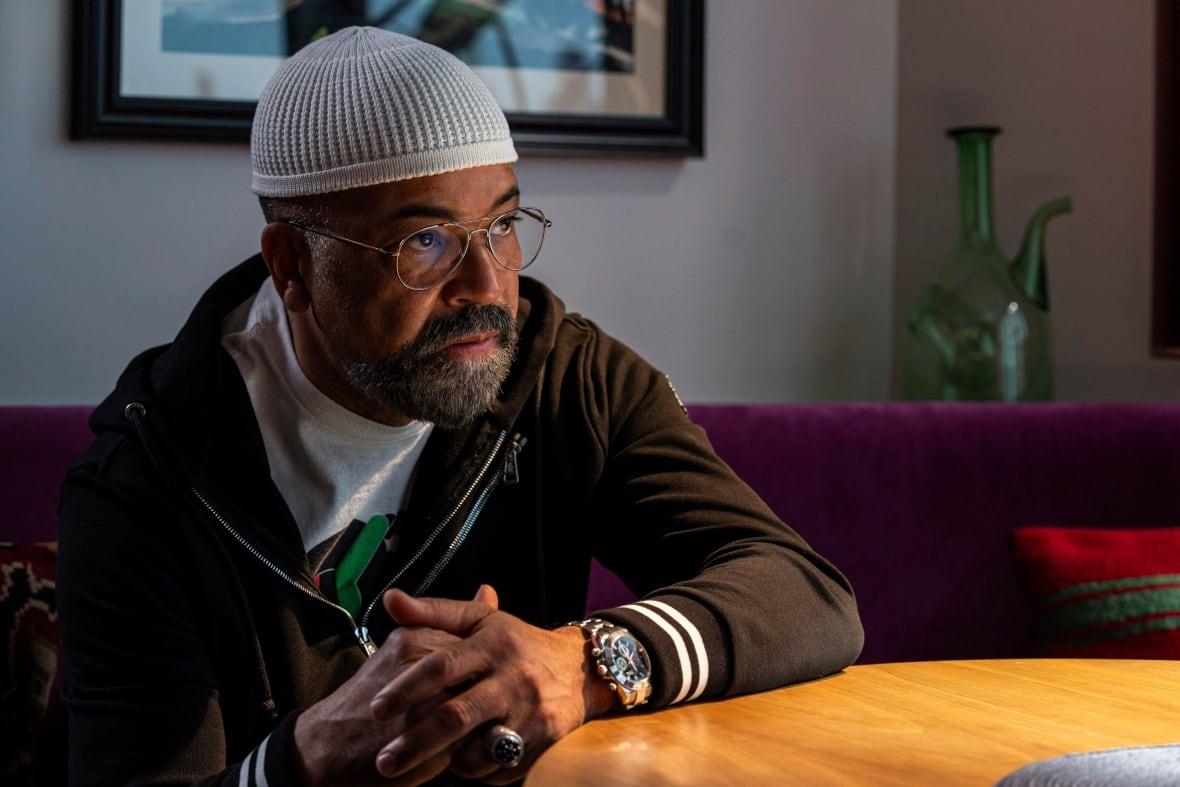 A man wearing a sweater and skullcap leans against a table.