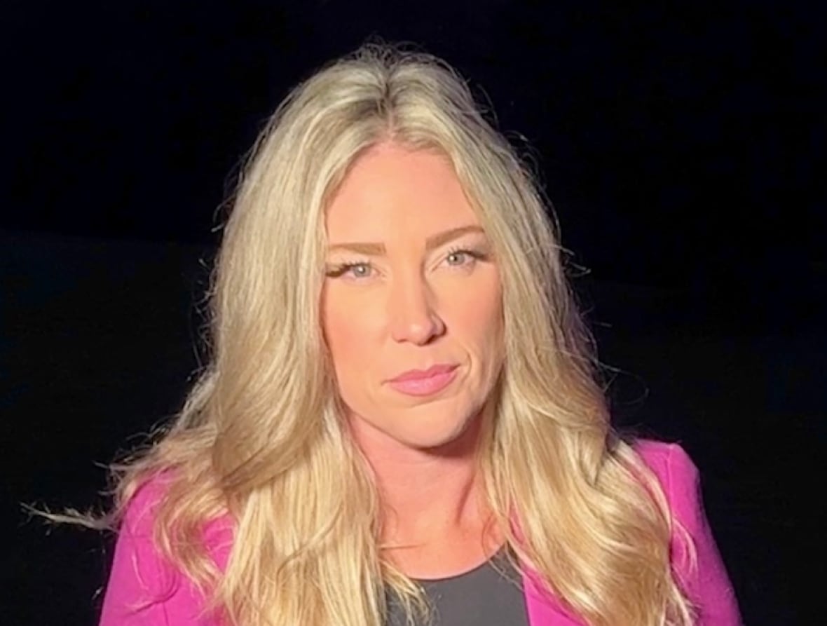 A woman with blond hair stands in front of a camera at sunset. She wears a pink blazer and black shirt.