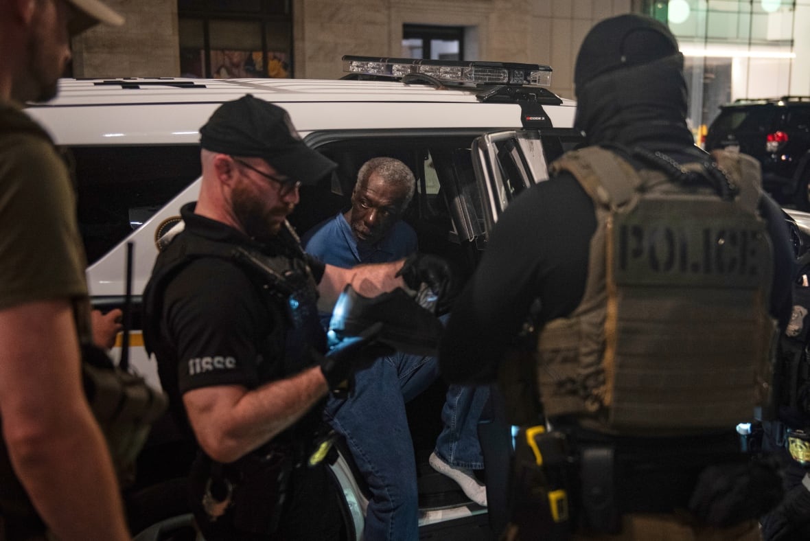 An older dark-complected man sits in the back of a police cruiser as two heavily armed men in uniforms stand nearby.