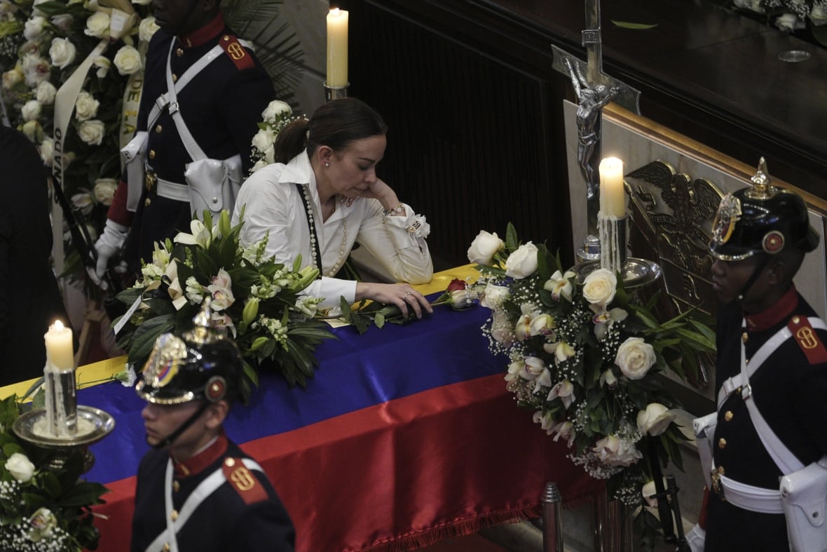 A woman leans over the coffin of her spouse, with three sentries in uniform standing nearby.