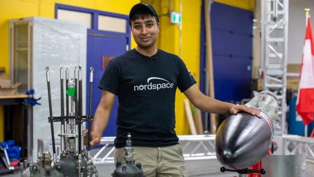 Rahul Goel, founder and CEO of NordSpace, stands in front of the company's Taiga rocket and some its engines which were 3D-printed in-house. All the rocket's components will be Canadian-made. A man in a black shirt and khaki pants stands between a rocket, on which one hand is placed, and rocket engines, on which the other hand is placed.